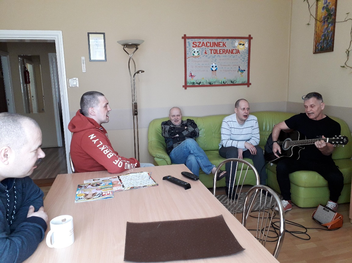 Grupa mężczyzn siedzących na kanapie jeden z nich trzyma w ręku gitarę.