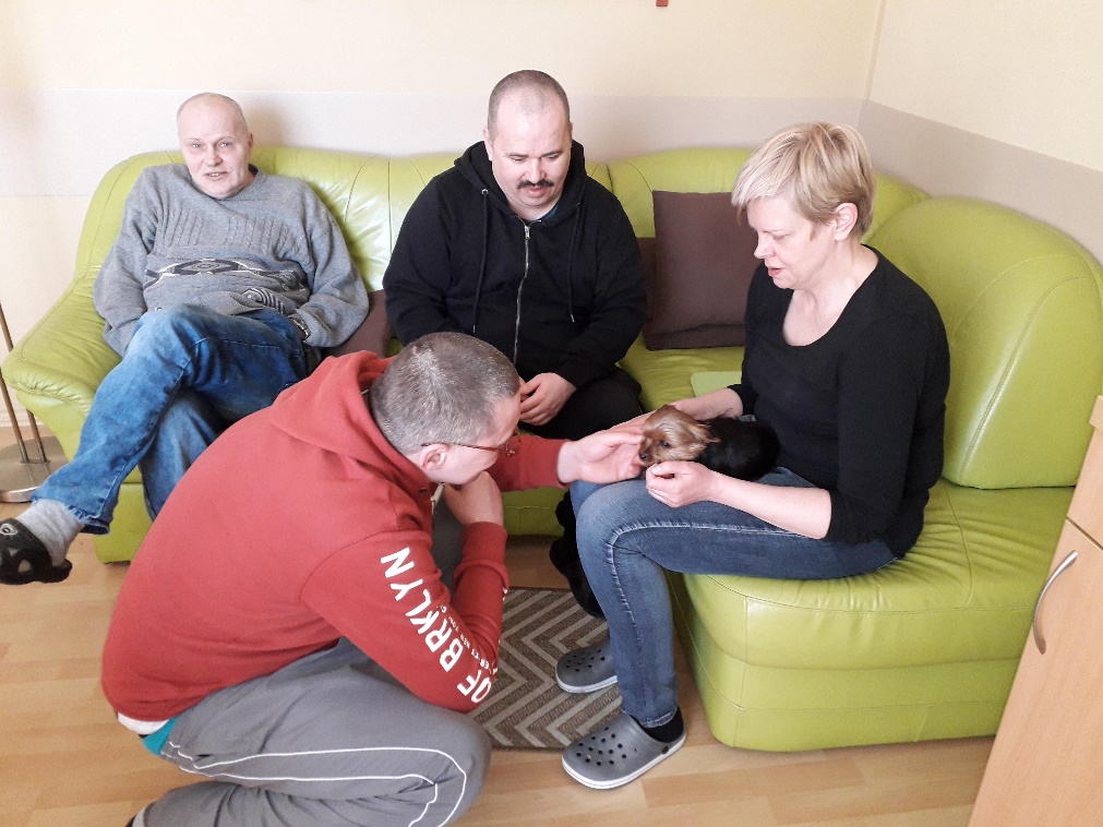 Grupa osób siedzących na kanapie, jedna z nich kobieta trzyma małego psa na kolanach.