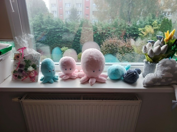 Na parapecie stoją pluszowe ośmiorniczki i inne miękkie dekoracje w pastelowych kolorach, obok bukiet materiałowych kwiatów i ozdobny pakunek.