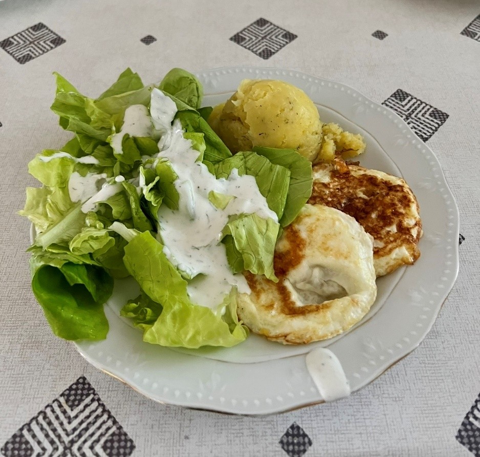Na białym talerzu leży porcja zielonej sałaty ze śmietaną, obok niej dwie sztuki sadzonych jajek oraz kulka gotowanych ziemniaków.