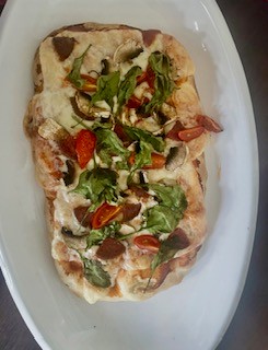 Prostokątna pizza na jasnym talerzu, z widocznymi składnikami: roztopionym serem, pomidorami koktajlowymi, pieczarkami, liśćmi rukoli i kawałkami kiełbasy.