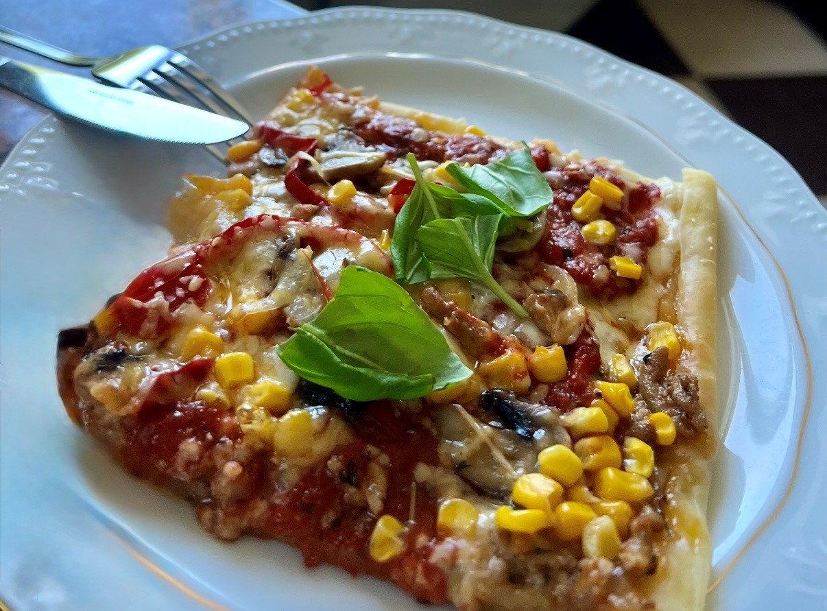 Na białym talerzu leży kwadratowy kawałek pizzy z żółtą kukurydzą, pieczarkami, sosem pomidorowym i listkami świeżej bazylii na wierzchu.