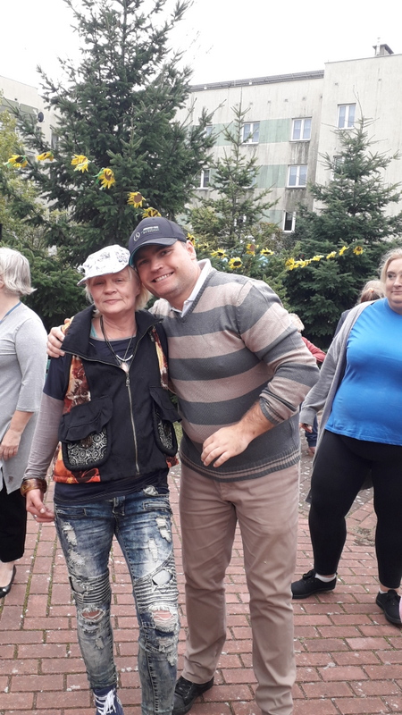 Kobieta i mężczyzna pozujący do zdjęcia na tle świerków z tyłu inne osoby spotkania.