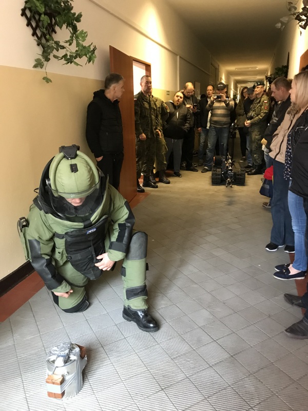 Trzech żołnierzy w jednostce wojskowej prezentują rozbrajanie ładunku wybuchowego, z tyłu kobiety i mężczyźni.