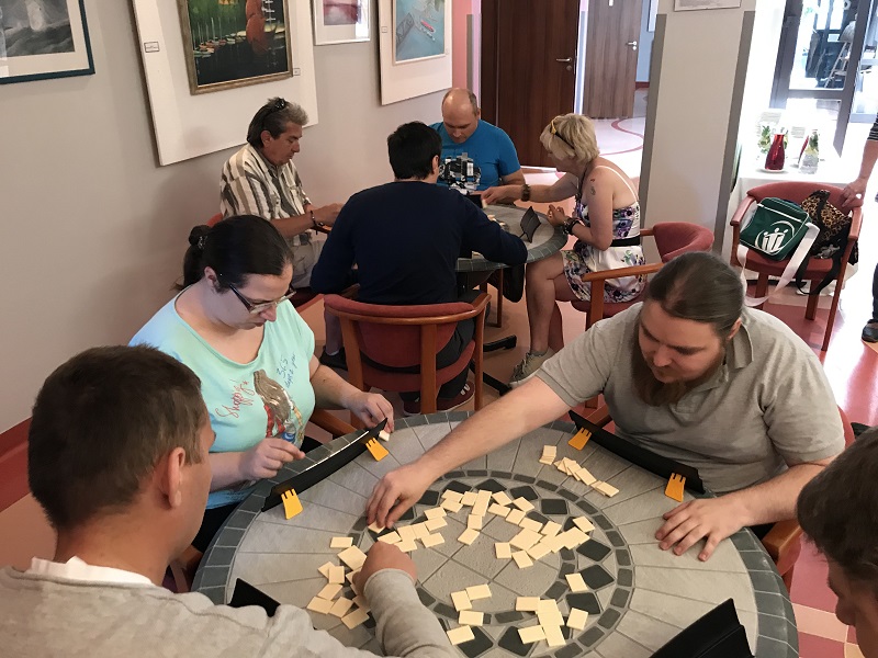 Dwa stoliki kawiarniane przy których siedzą kobiety i mężczyźni rozgrywający grę rummikub. 