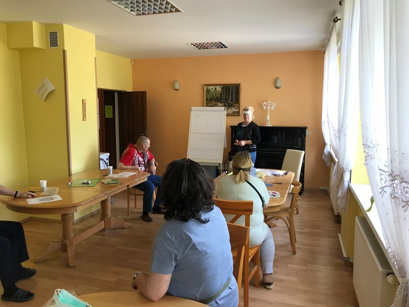 Czterech uczestników przy stołach oraz terapeuta prowadzący zajęcia terapeutyczne. 
