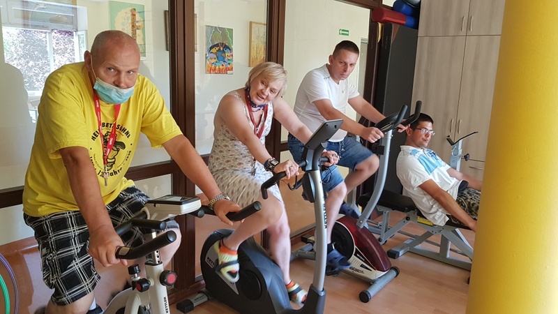 Trzech mężczyzn i kobieta ćwiczą na sprzętach stacjonarnych znajdujących się w sali sportowej.