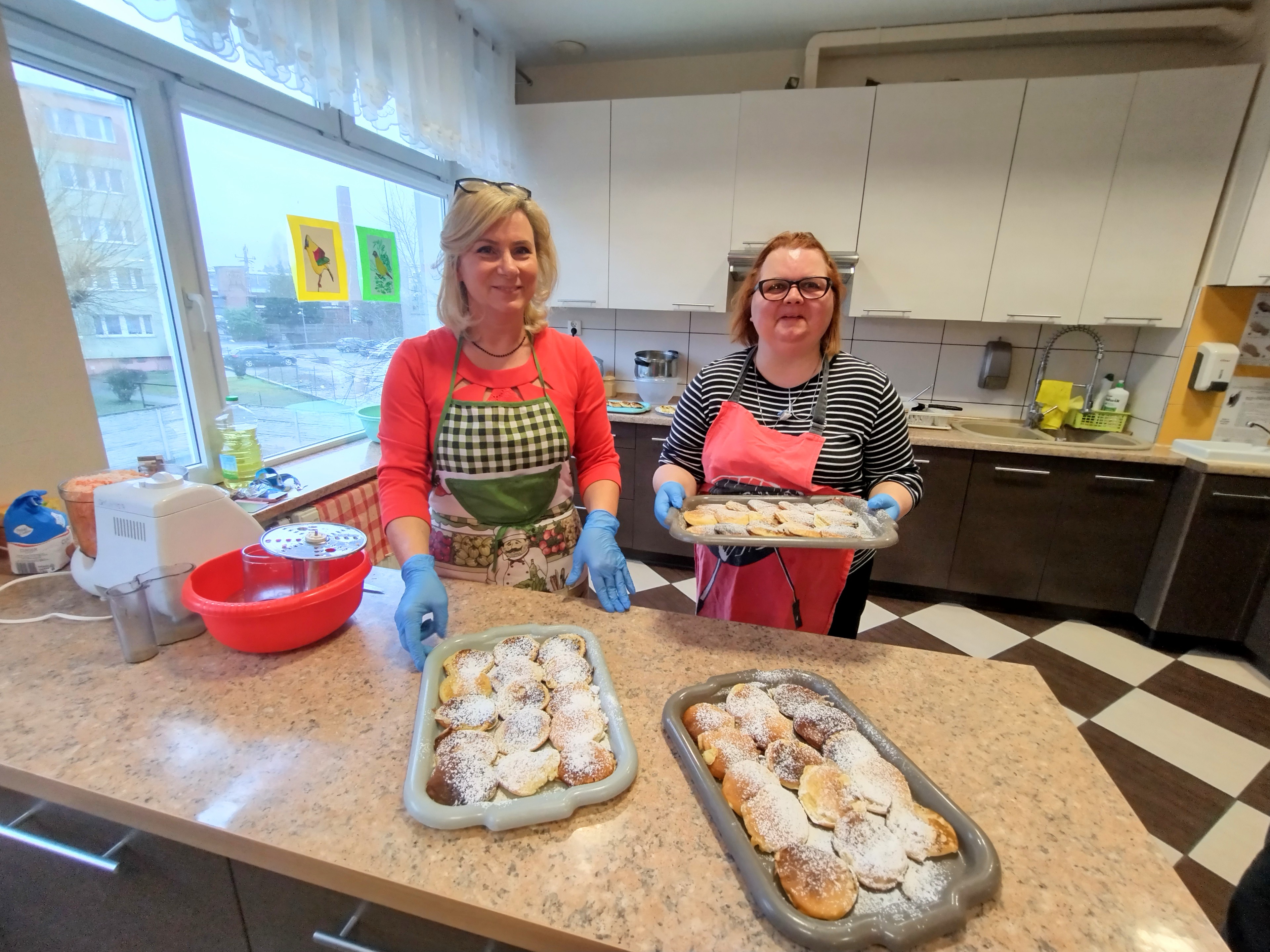 Na kuchennym blacie leżą dwie tace z plackami, za blatem stoją dwie kobiety jedna z nich trzyma tacę z plackami. 