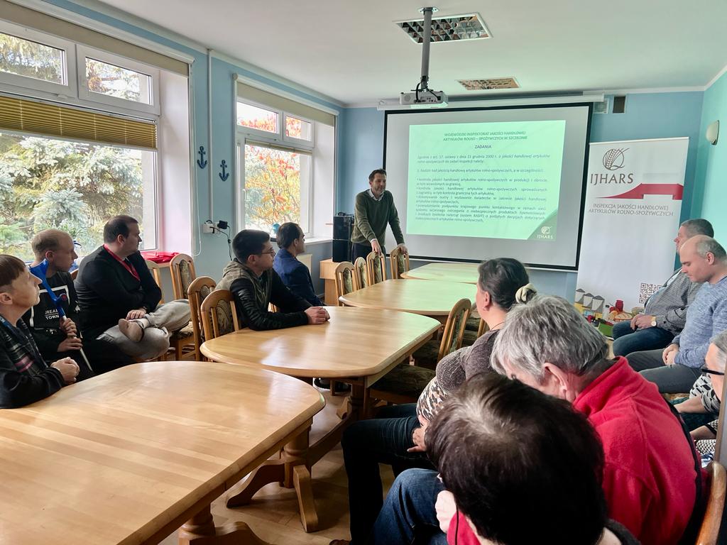 Na pierwszym planie stoły przy których siedzą kobiety i mężczyźni słuchający prelekcji. W tle mężczyzna zwrócony twarzą do grupy, za nim ekran wyświetlający obraz z rzutnika.