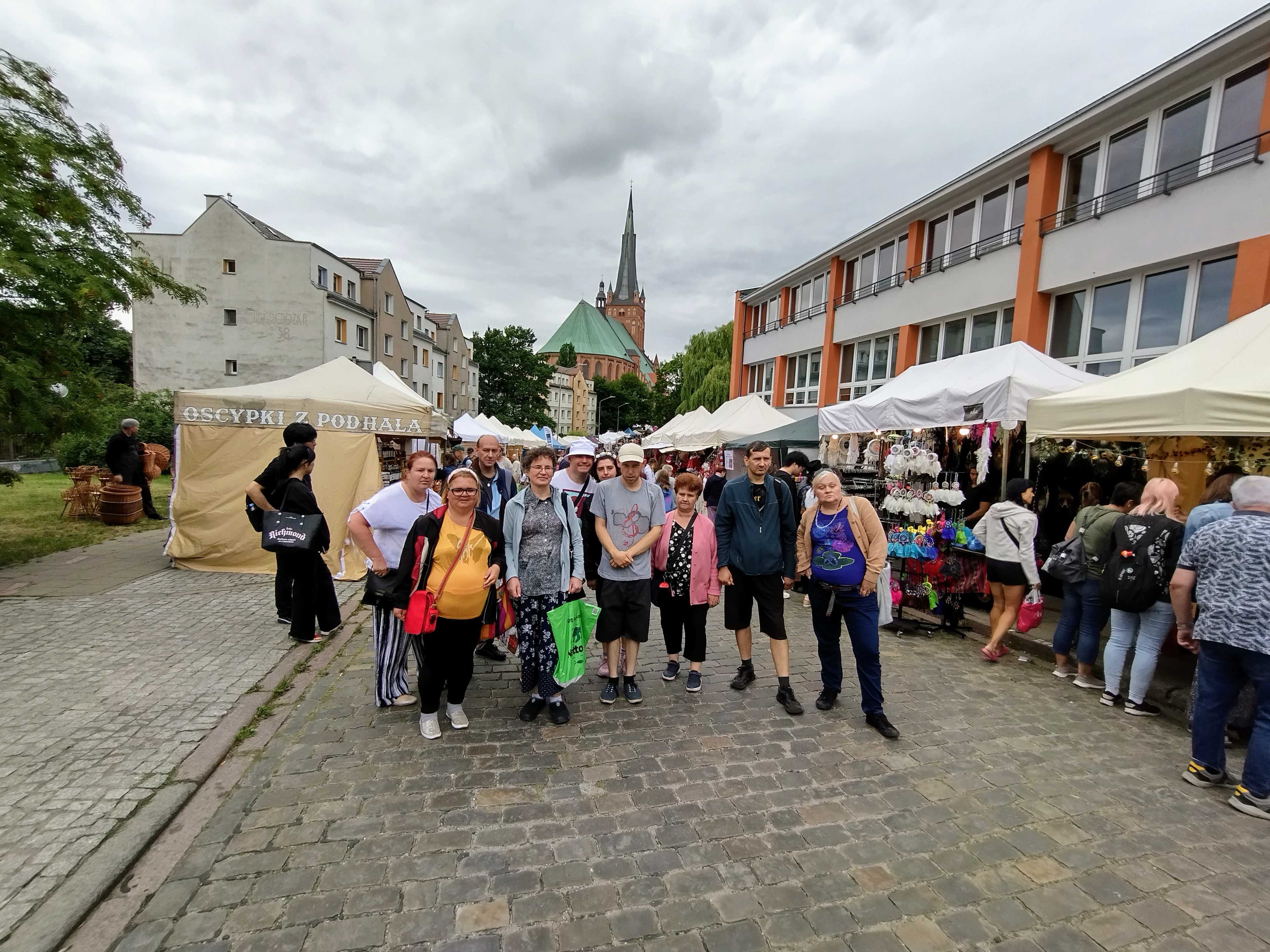 Grupa kobiet i mężczyzn stoi na brukowanej ulicy. W tle stoiska wystawców i budynek katedry.