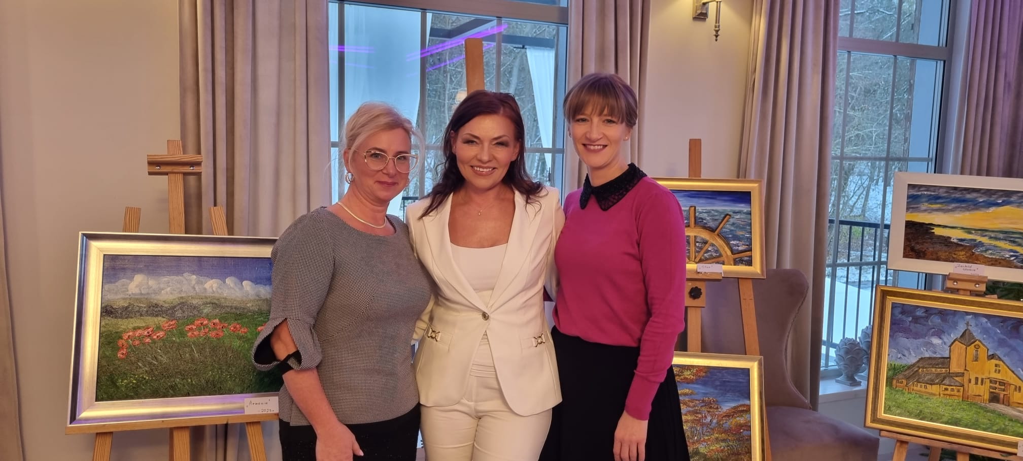 Trzy uśmiechnięte i elegancko ubrane kobiety stoją blisko siebie. Na drugim planie wiele prac malarskich stoi na sztalugach. 