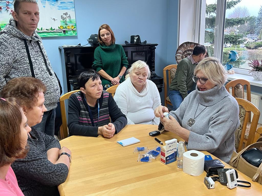 Przy stole siedzą kobiety. Na blacie leży glukometr i jednorazowe igiełki. Kobieta po prawej otwiera paski do pomiaru poziomu cukru.