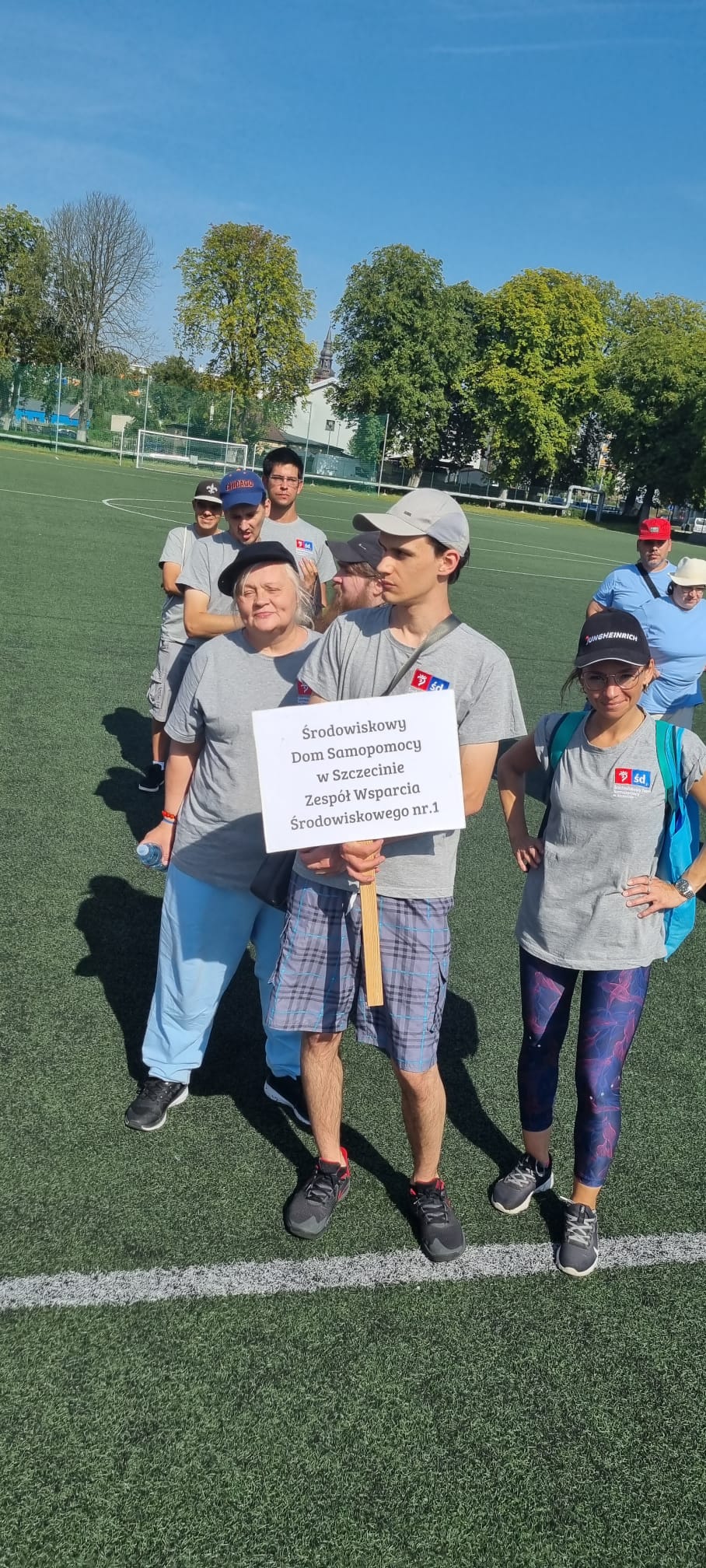 Na zielonej murawie boiska stoi siedem osób jeden za drugim. 