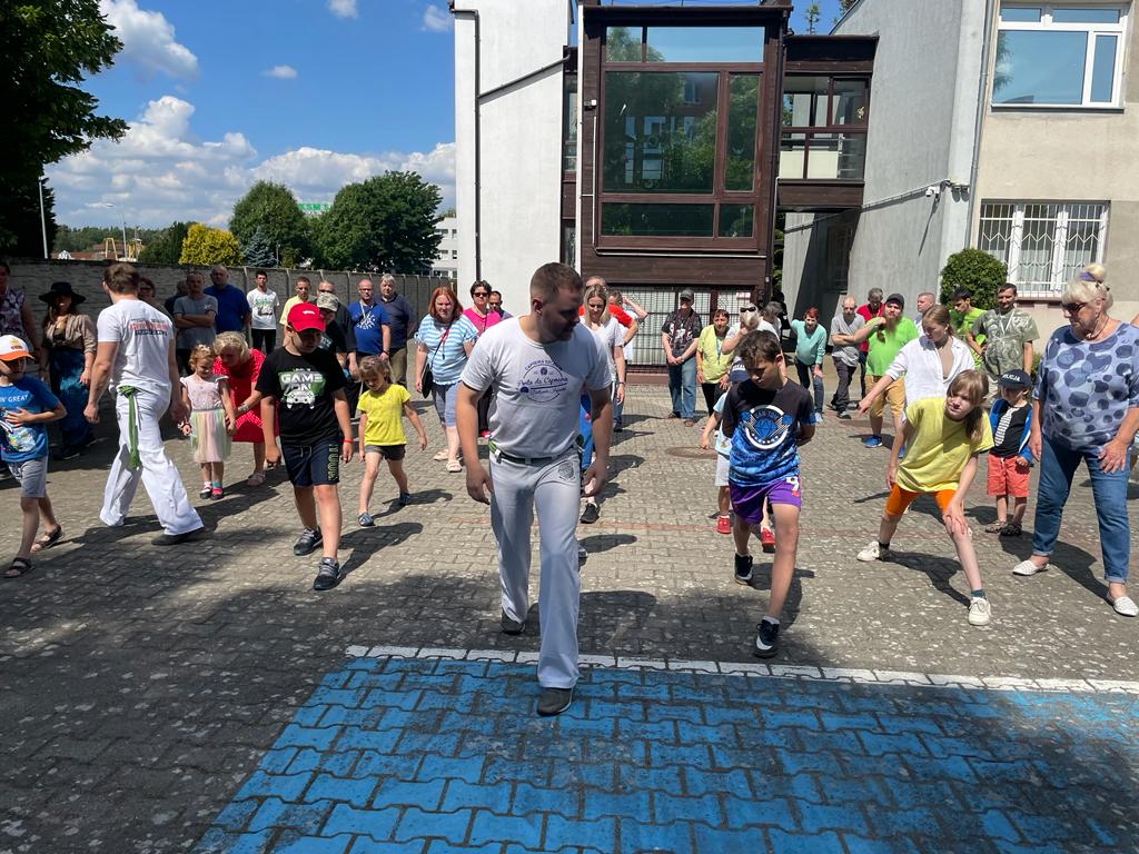 Grupa kobiet, mężczyzn i dzieci na parkingu przed ośrodkiem wykonuje ćwiczenia naśladując trenera. 