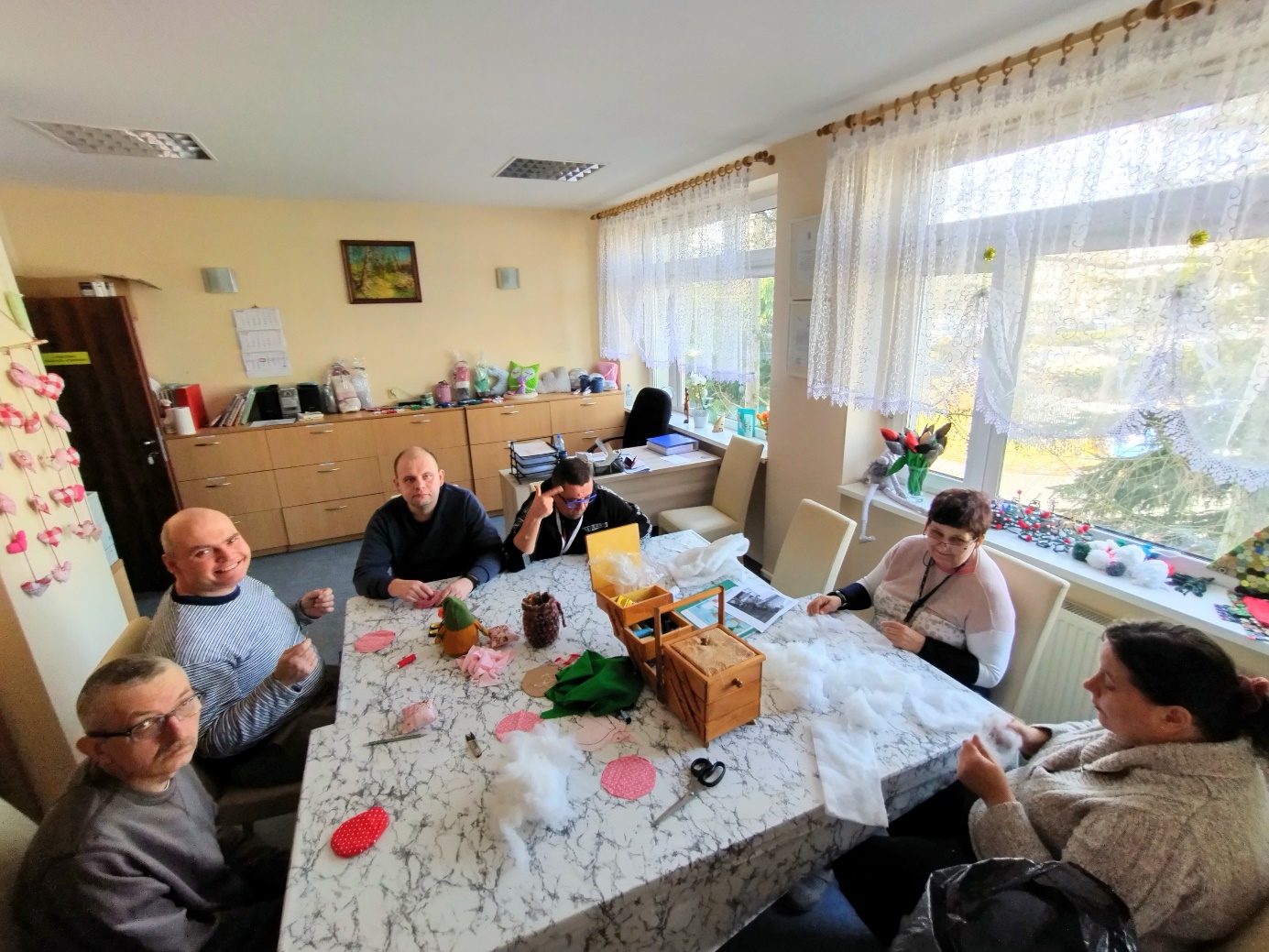 Dwie kobiety i czterech mężczyzn siedzi  przy stole szyjąc lub segregując materiały krawieckie