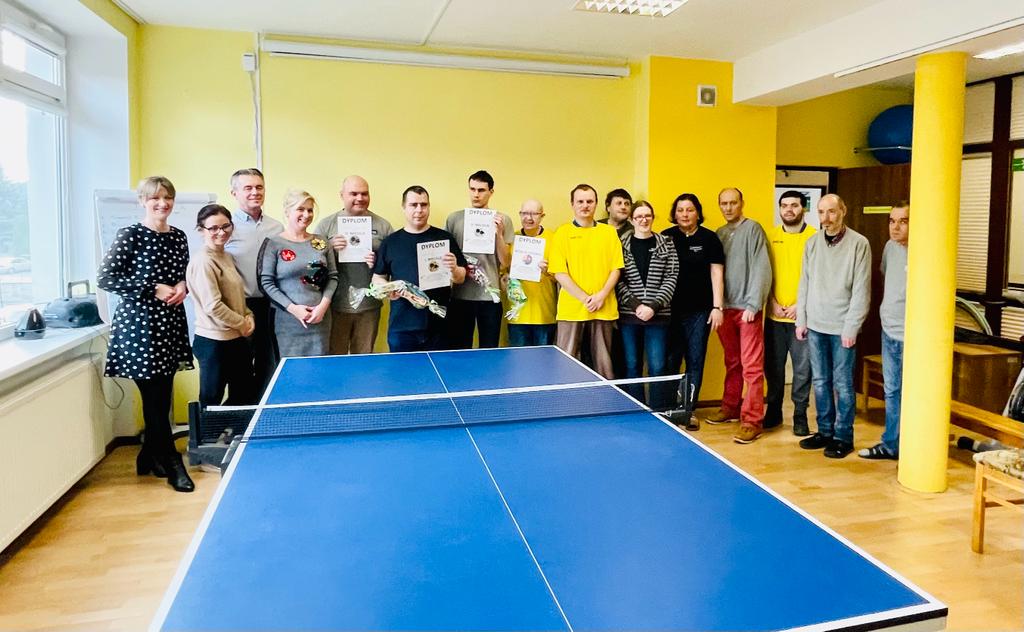 Grupa kobiet i mężczyzn stojących za stołem tenisowym. Czterech mężczyzn trzyma w rękach dyplomy oraz  nagrody za udział w konkursie.