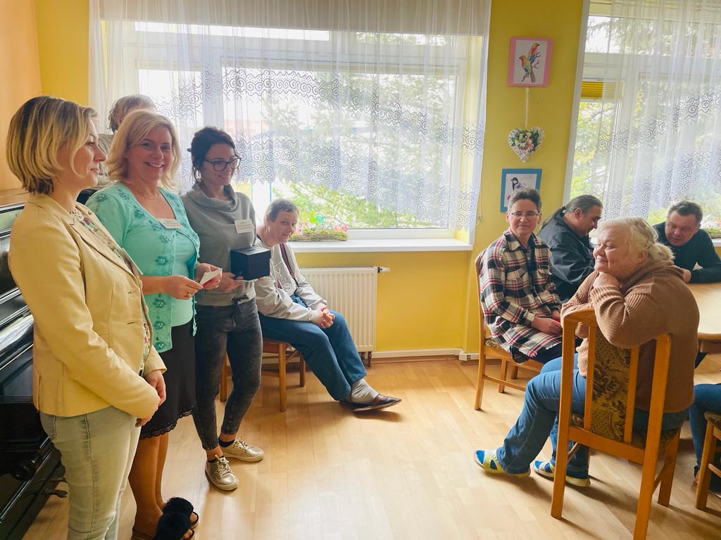 Po lewej stronie stoją trzy kobiety z komisji wyborczej, jedna z nich trzyma urnę. Po prawej grupa kobiet i mężczyzn siedzących na krzesłach.