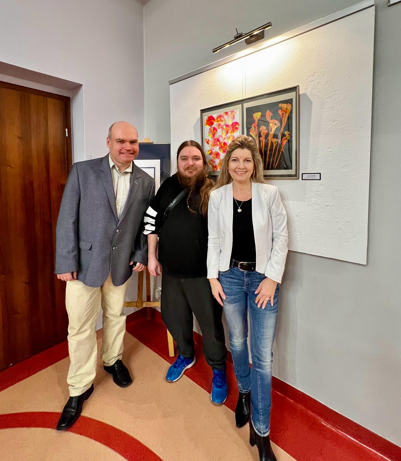 Dwóch mężczyzn i kobieta stoją uśmiechnięci i pozują do zdjęcia na tle obrazów. 