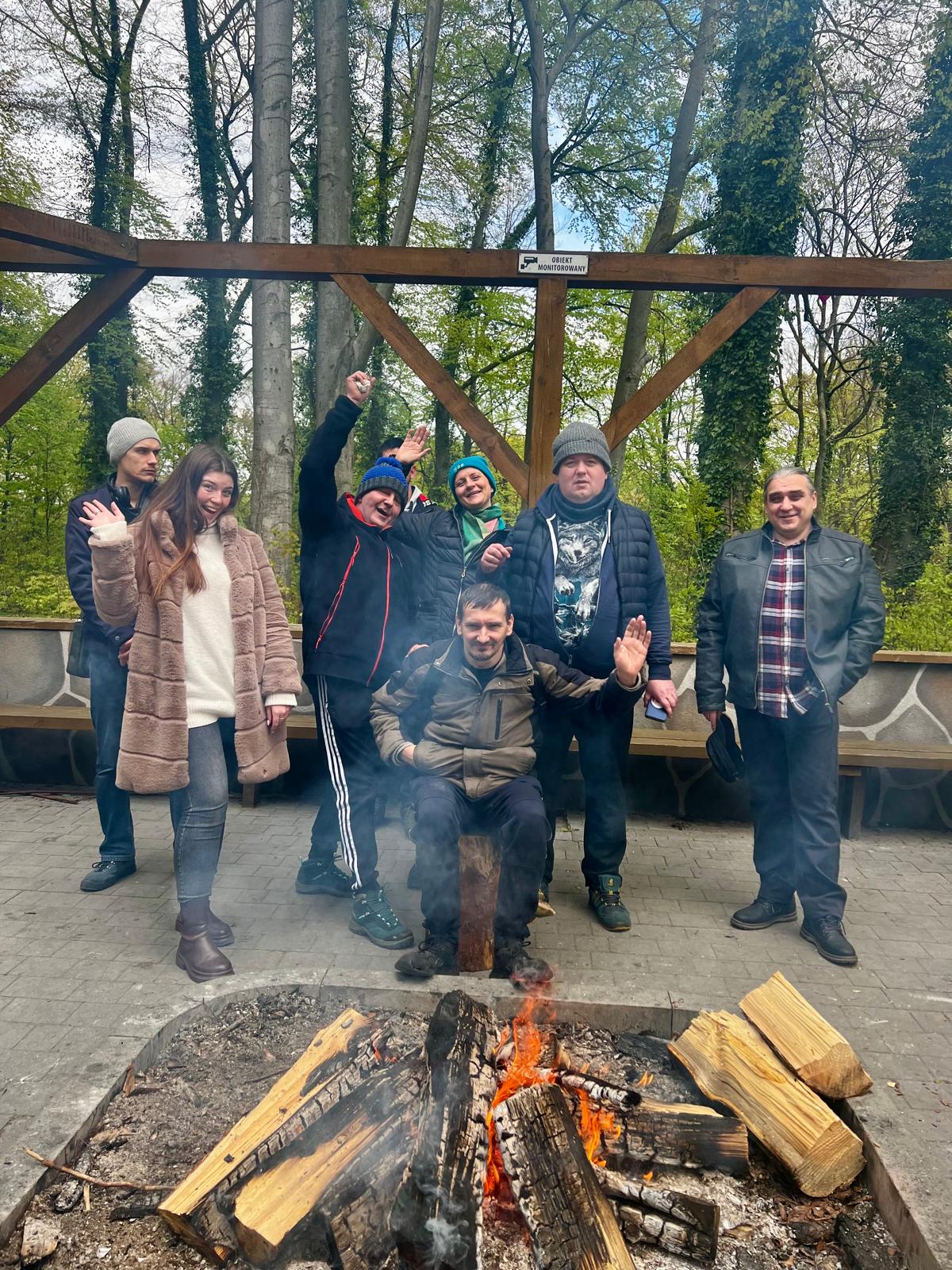Na pierwszym planie ognisko a za nim grupa kobiet i mężczyzn uśmiechają się i machają do obiektywu.