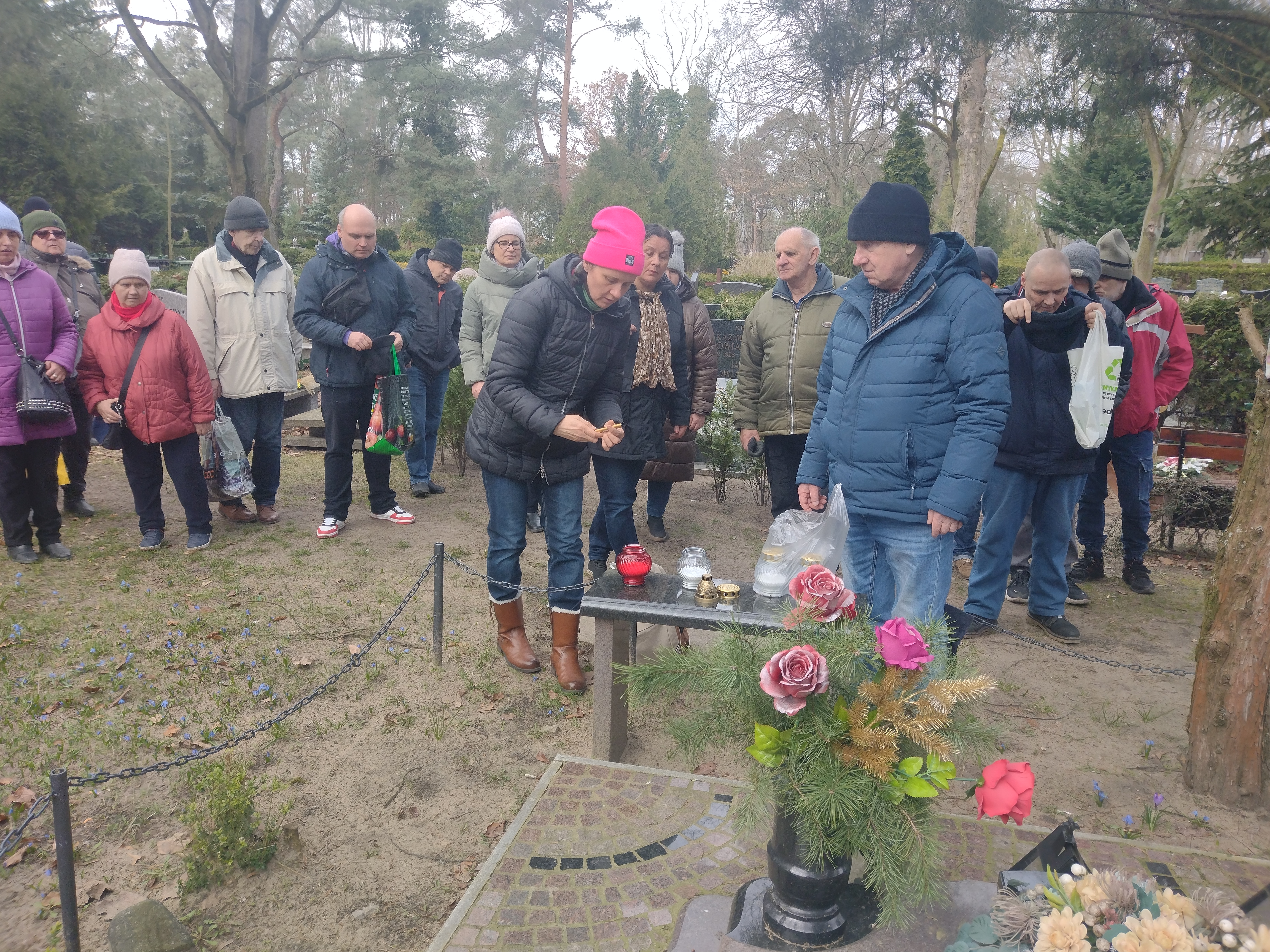 Przy cmentarnej ławeczce kobieta w czapce podpala znicz. W tle grupa osób pogrążonych w zadumie. 