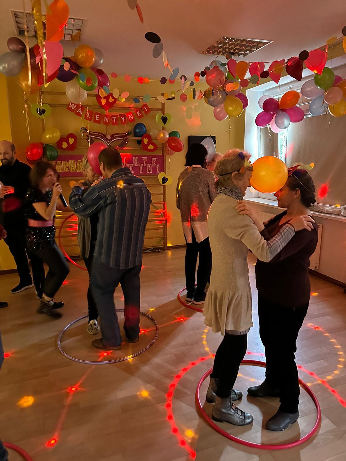 Na pierwszym planie kobieta i mężczyzna tańczą trzymając się za ręce. Na drugim planie wiele osób tańczących. Pod sufitem kolorowe girlandy i balony.   
