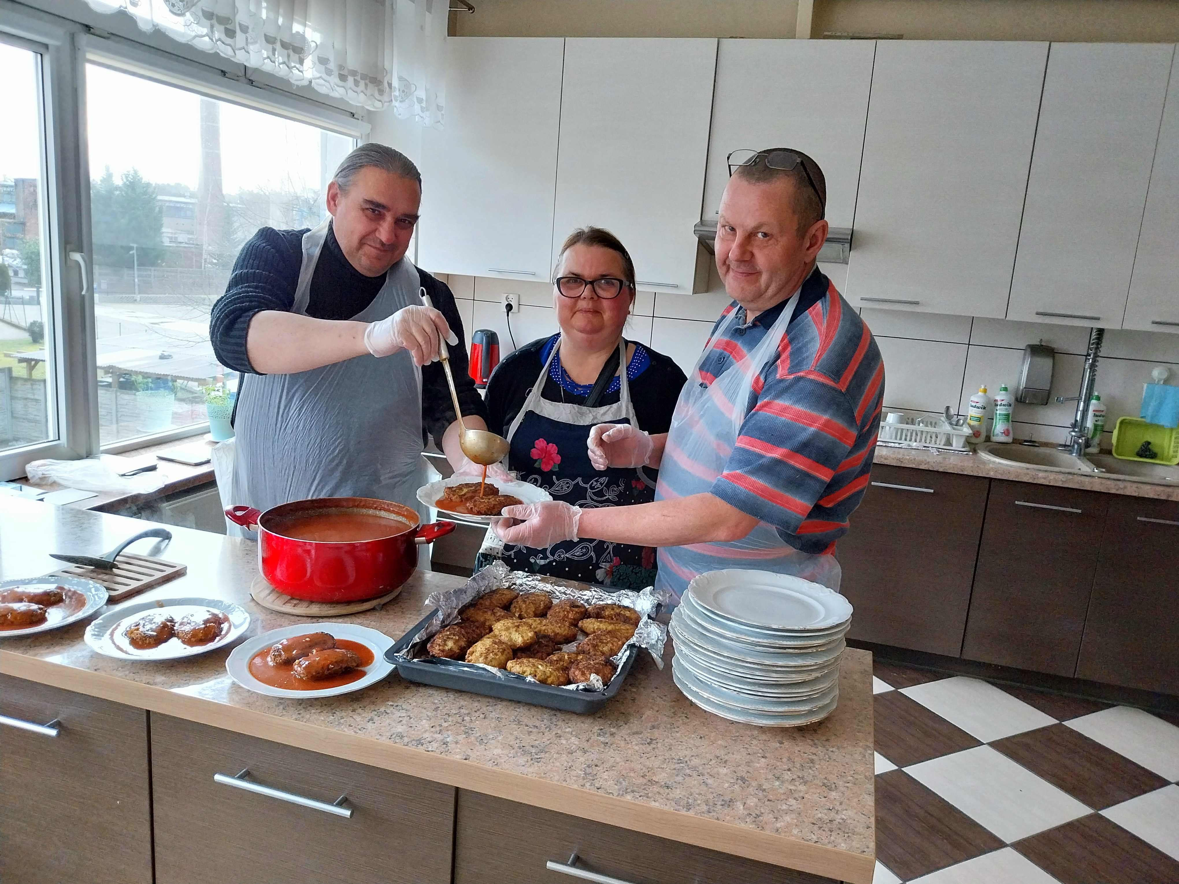 Na pierwszym planie blat kuchenny, na którym leżą talerze z nałożonymi gołąbkami polanymi sosem pomidorowym. Na drugim planie 3 uśmiechnięte osoby nakładają gołąbki na talerze i polewają je sosem.