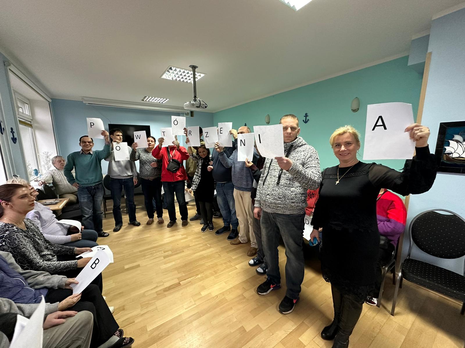 Na sali grupa osób ustawiona w szeregu. Każda osoba trzyma kartkę z literą. Litery układają się w napis: POWTÓRZENIA.