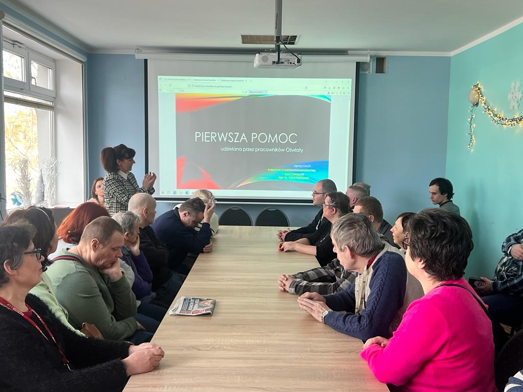 Na ekranie napis : pierwsza pomoc, przed ekranem stoi kobieta zwrócona w prawą stronę  do osób siedzących przy stole. 