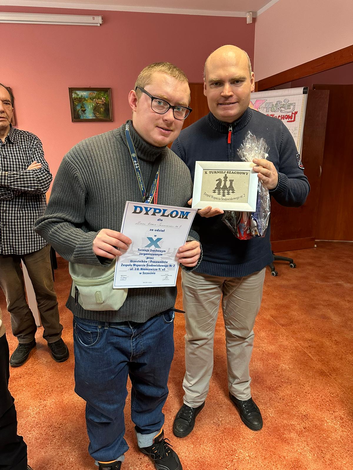 Dwóch uśmiechniętych mężczyzn trzyma w rękach dyplom i nagrodę za udział w turnieju. 