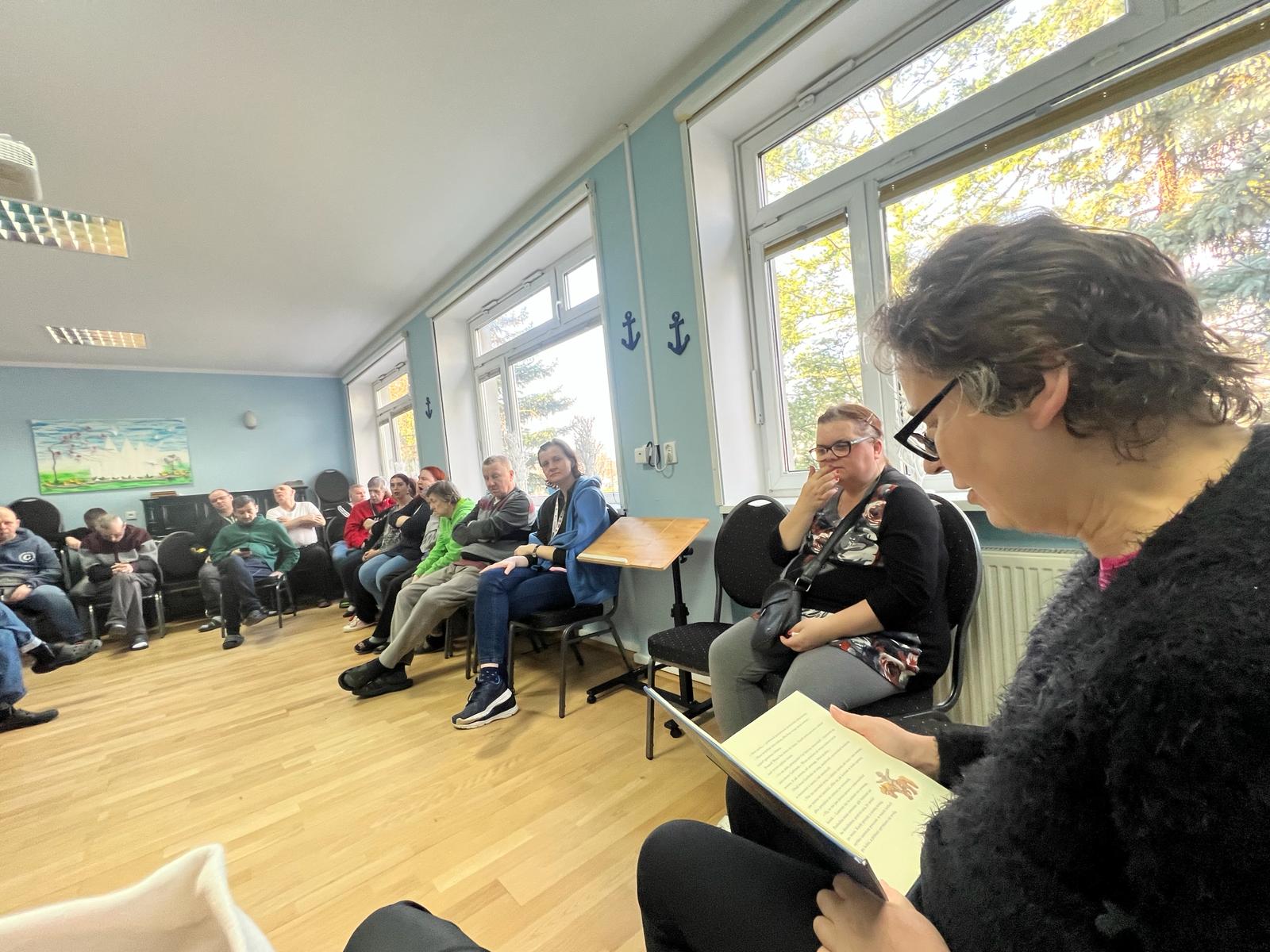 Po prawej stronie siedzi kobieta w okularach i czyta książkę. Na drugim planie pod ścianą siedzą pozostałe osoby i słuchają. 