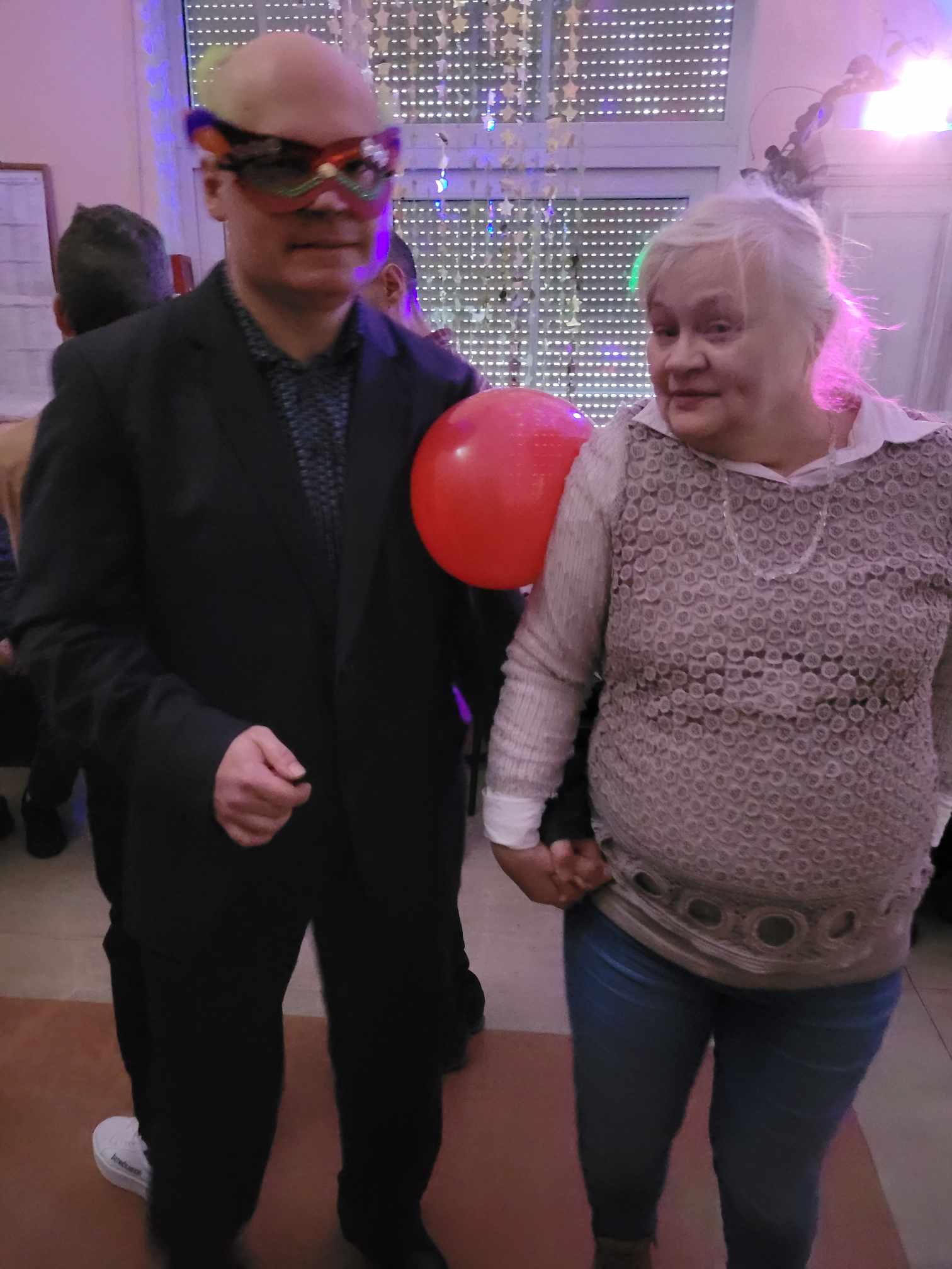 Po lewej stoi mężczyzna w masce, po prawej stoi kobieta a miedzy ich ramionami czerwony balon.