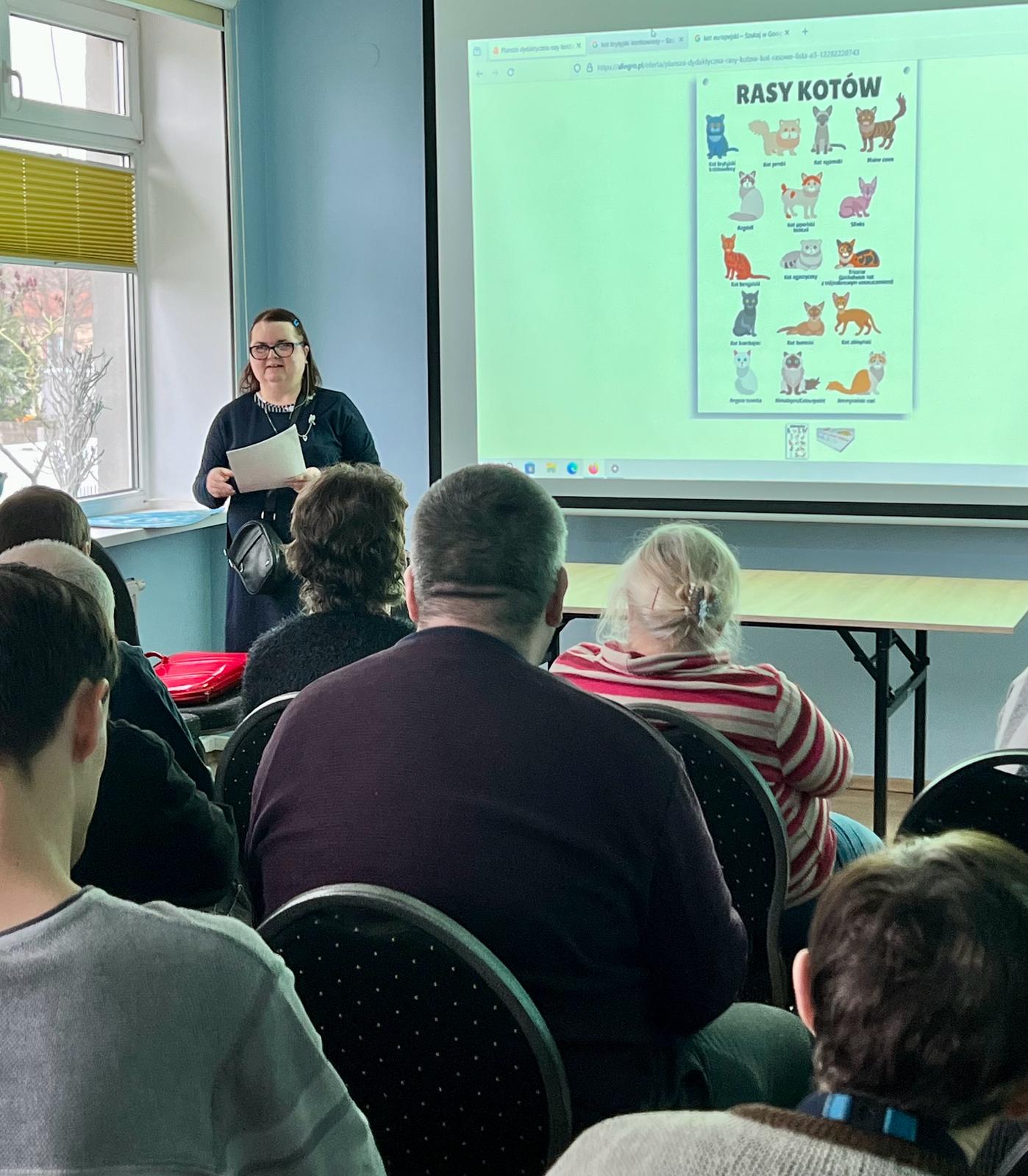 Na pierwszym planie tyłem siedzą osoby słuchające prelekcji. Na drugim planie po lewej kobieta z kartką w ręku, po prawej ekran z obrazkami kotów.