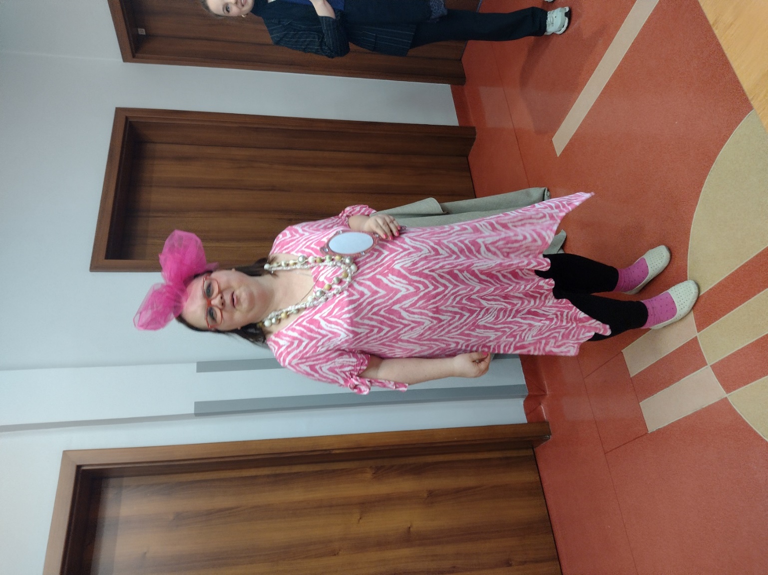Kobieta w różowej sukience i różowej kokardzie na głowie stoi uśmiechnięta i trzyma w ręku lusterko. 