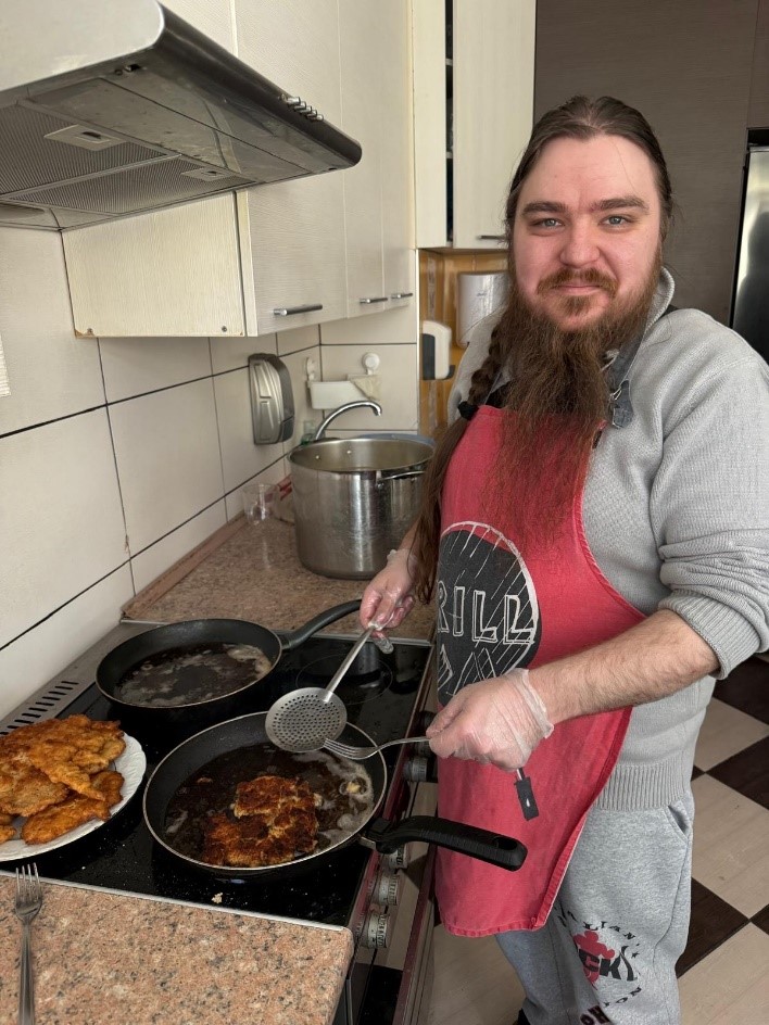Mężczyzna z brodą trzyma widelec i łopatkę  smażąc kotlety. 