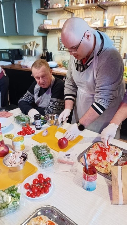 Na zdjęciu jest dwóch mężczyzn. Jeden mężczyzna kroi warzywa. Na stole leżą warzywa. 