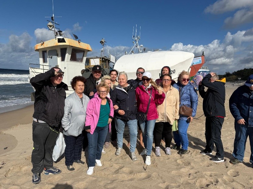 Kilkunastu osobowa grupa w słoneczny dzień stoi na plaży i pozuje do zdjęcia. W tle biały kuter rybacki.