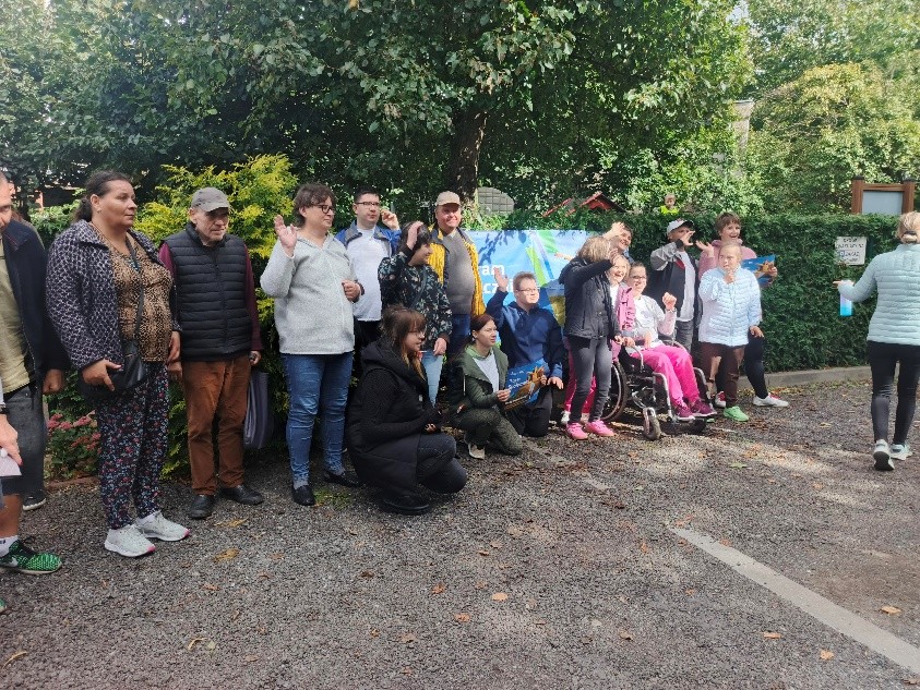 Duża grupa osób stoi i pozuje do zdjęcia. Jedna osoba jest na wózku inwalidzkim. W tle zielone korony drzew. 