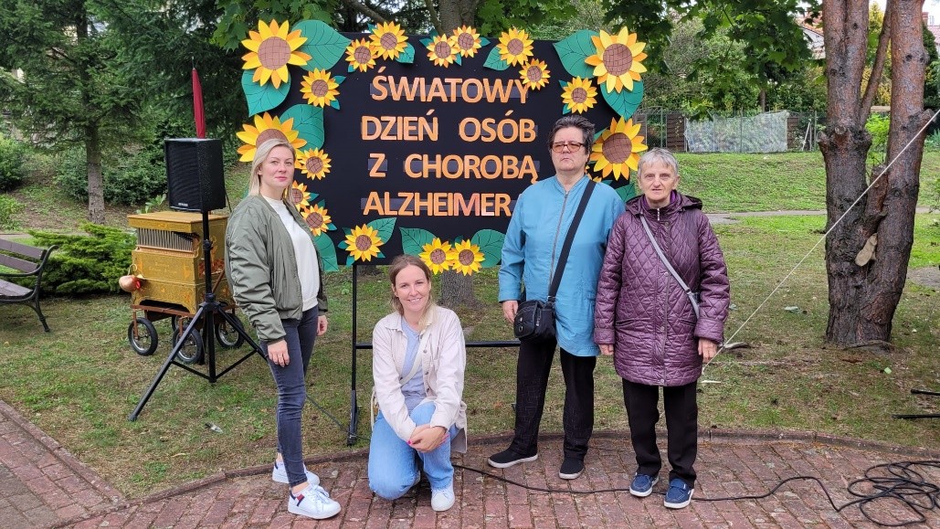 Cztery kobity pozują do zdjęcia na tle tablicy z napisem „ Światowy Dzień Osób z Chorobą Alzheimera”.