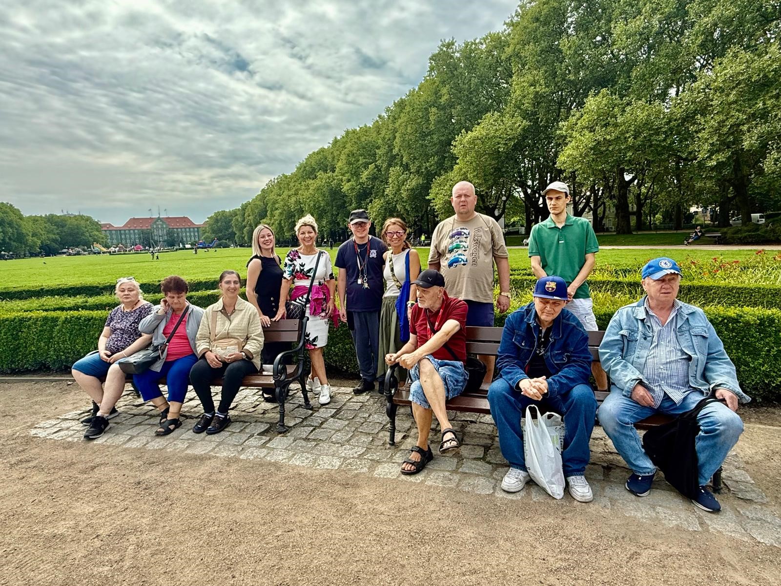 : Grupa osób siedzi na dwóch ławkach. Między ławkami stoi kilka osób. W tle widok na Urząd Miasta i Jasne Błonia.