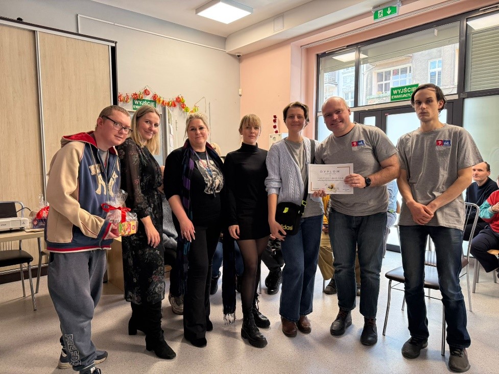 Kilka osób stoi w sali przy dużych oknach, trzymając dyplom i kolorowe paczki.