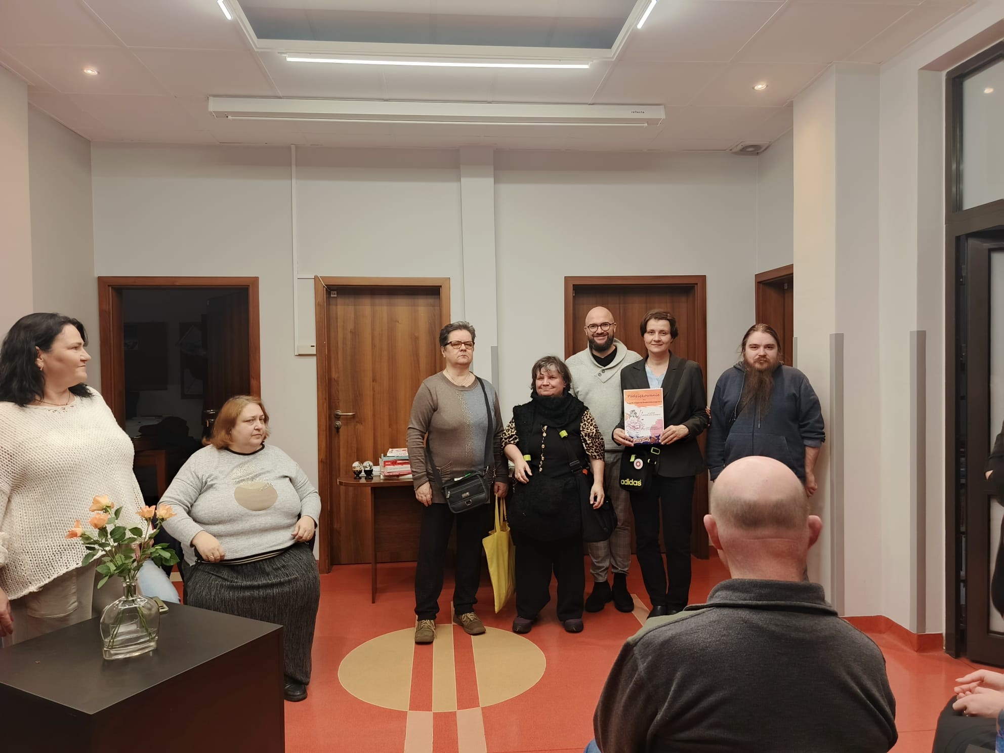 Grupa osób stoi w jasnym pomieszczeniu z czerwono-beżową podłogą, w tle drzwi i stolik z książkami.