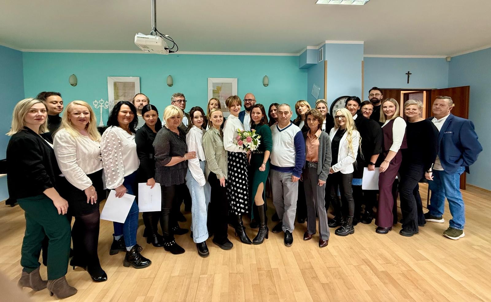 Duża grupa osób stoi w jasnej sali z turkusową ścianą, drewnianą podłogą i bukietem kwiatów na pierwszym planie.