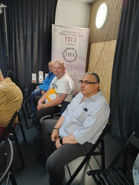 W ciemnej sali na krzesłach siedzi 3 mężczyzn. W tle plakat z napisem TRUE.