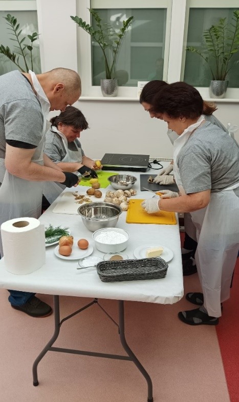 Wokół stołu stoją cztery osoby i kroją warzywa na deskach. Na stole stoi kuchenka indukcyjna, ziemniaki, pieczarki i jajka.