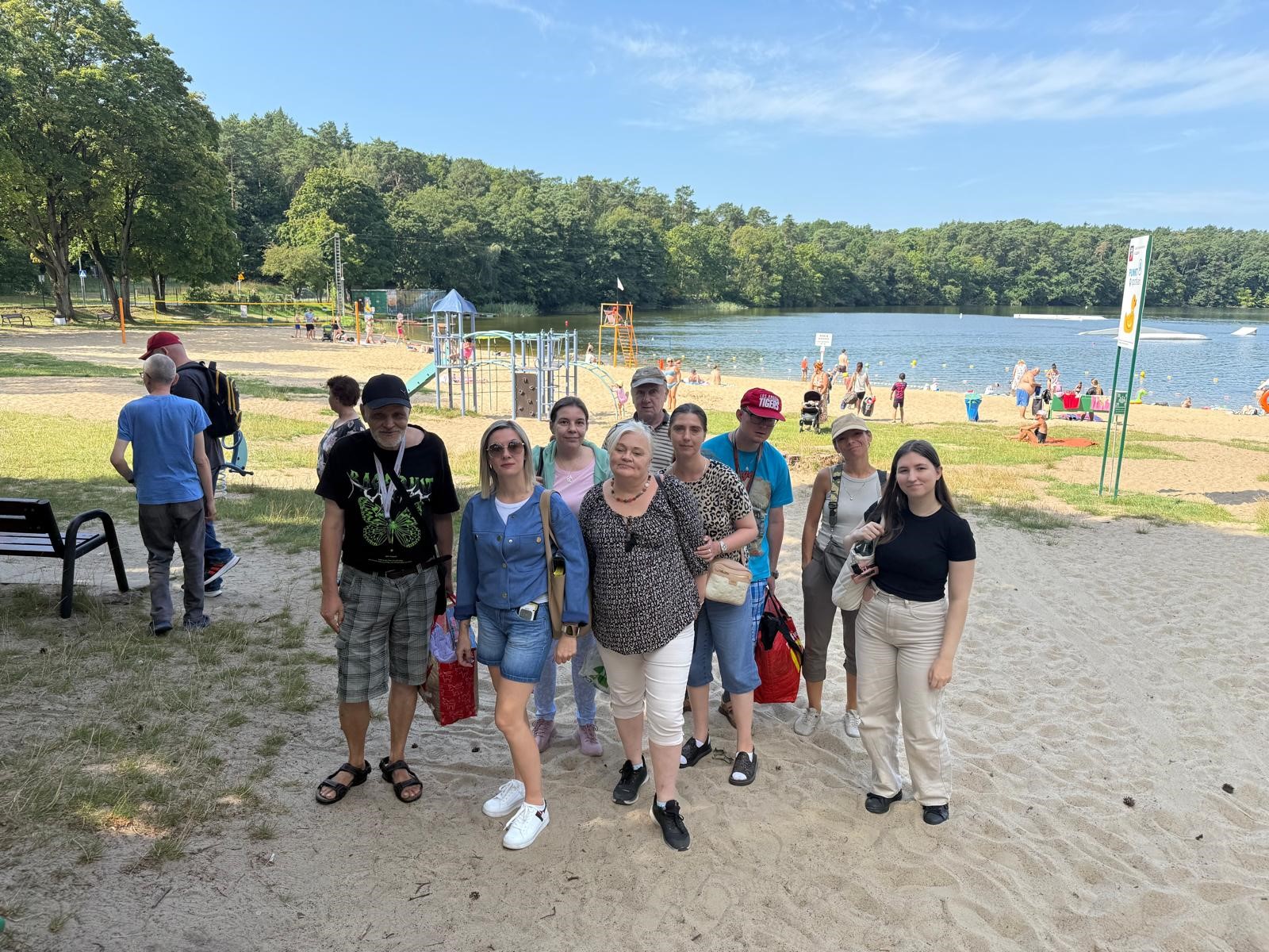 Na pierwszym planie stoi grupa ludzi pozująca do zdjęcia na plaży. W tle jezioro, las i błękitne niebo.