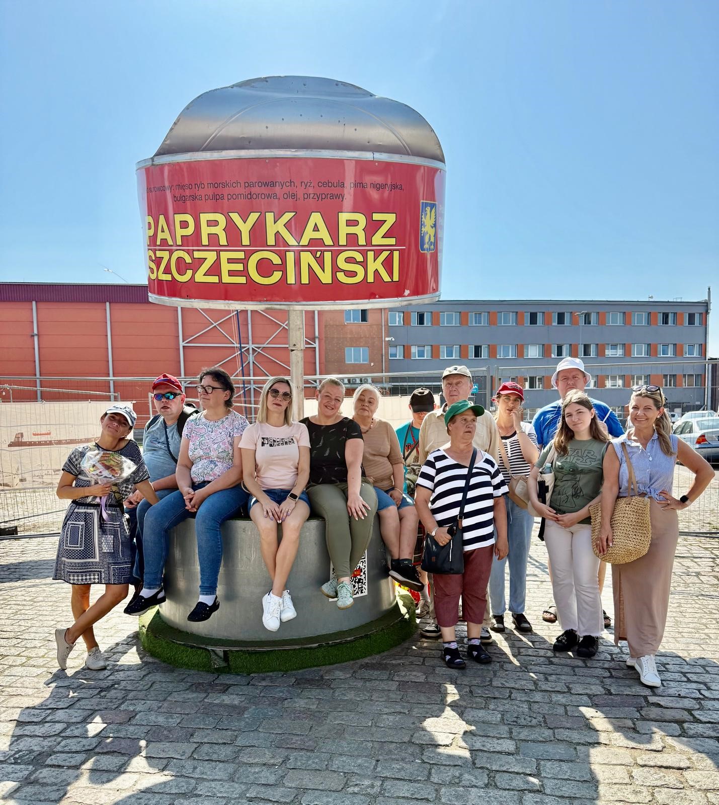 Na tle niebieskiego słonecznego nieba pomnik Paprykarza Szczecińskiego, pod którym pozuje grupa wielu osób.