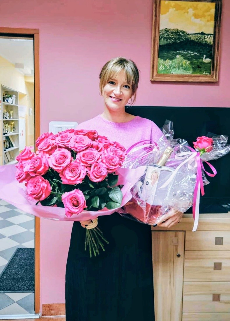 Uśmiechnięta kobieta patrzy w obiektyw stojąc i trzymając wielki bukiet różowych róż oraz prezent zawinięty w celofan.