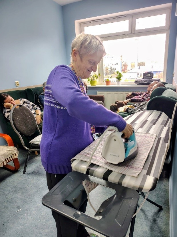 Osoba w filetowym swetrze prasuje materiał na desce w pomieszczeniu z niebieskimi ścianami. W tle widać okno z doniczkami i kilka osób siedzących na kanapach.