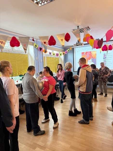 Grupa osób tańczy i bawi się w udekorowanej Sali. Przy suficie girlandy z serc i balonów. Atmosfera jest radosna i towarzyska.