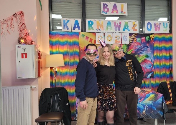 Troje osób stoi razem na tle kolorowej, tęczowej dekoracji z napisem ‘Bal Karnawałowy 2026’. Jedna z osób ma na głowie imprezowy stożkowy kapelusz. Wokół widać ozdoby i elementy wystroju sali.
